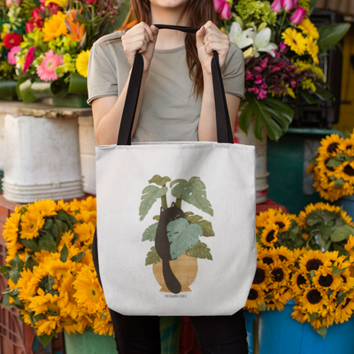 A woman holding up a beige cat tote bag with black handles featuring a black cat munching on monstera plant leaves.